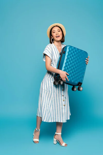 Sorrindo morena asiática menina em vestido listrado e chapéu de palha com saco de viagem no fundo azul — Fotografia de Stock