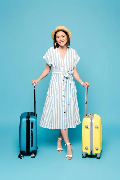 Lächelnde brünette asiatische Mädchen in gestreiftem Kleid und Strohhut mit Reisetaschen auf blauem Hintergrund — Stockfoto