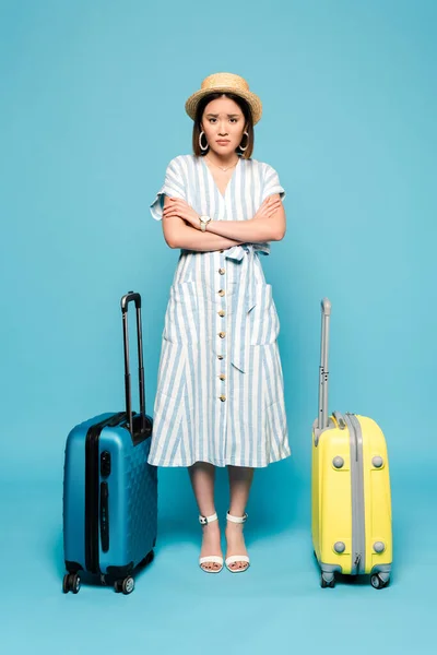 Morena triste chica asiática en vestido a rayas y sombrero de paja con bolsas de viaje sobre fondo azul - foto de stock