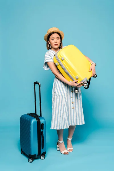 Morena triste chica asiática en vestido a rayas y sombrero de paja con bolsas de viaje sobre fondo azul - foto de stock