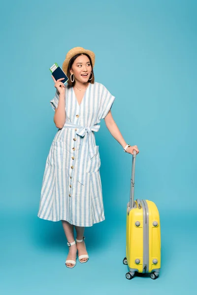 Smiling brunette asian girl in striped dress and straw hat with travel bag and passport with air ticket on blue background — Stock Photo