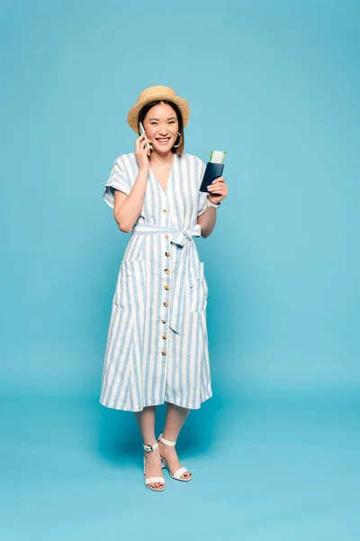 Улыбающаяся брюнетка азиатка в полосатом платье и соломенной шляпе с паспортом и авиабилетом разговаривает по смартфону на синем фоне — стоковое фото