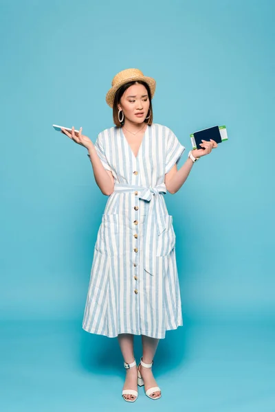 Sad brunette asian girl in striped dress and straw hat with passport, air ticket, smartphone on blue background — Stock Photo