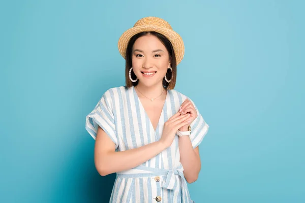 Sonriente morena asiática chica en vestido a rayas y sombrero de paja sobre fondo azul - foto de stock
