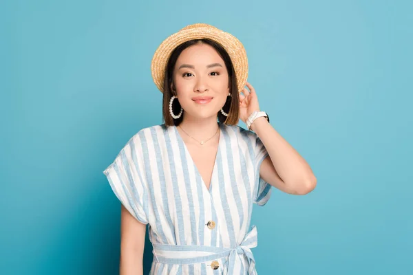 Sonriente morena asiática chica en vestido a rayas y sombrero de paja sobre fondo azul - foto de stock
