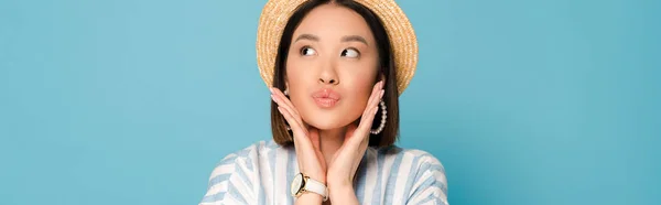 Brunette asian girl in striped dress and straw hat touching face and pouting lips on blue background, panoramic shot — Stock Photo