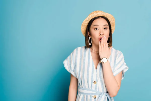 Impactado morena chica asiática en vestido a rayas y sombrero de paja que cubre la boca con la mano sobre fondo azul - foto de stock