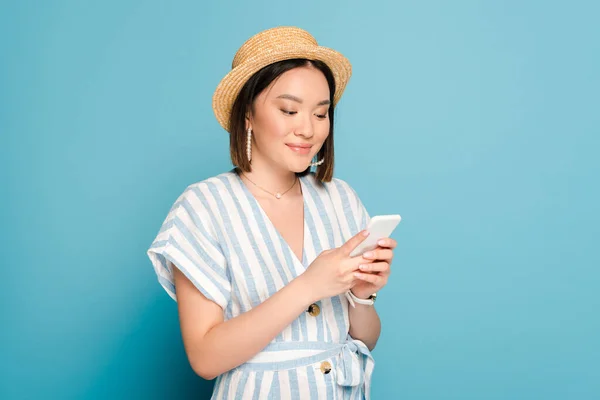 Усміхнена брюнетка азіатська дівчина в смугастій сукні і солом'яному капелюсі використовує смартфон на синьому фоні — стокове фото