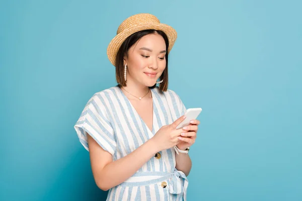 Smiling brunette asian girl in striped dress and straw hat using smartphone on blue background — Stock Photo