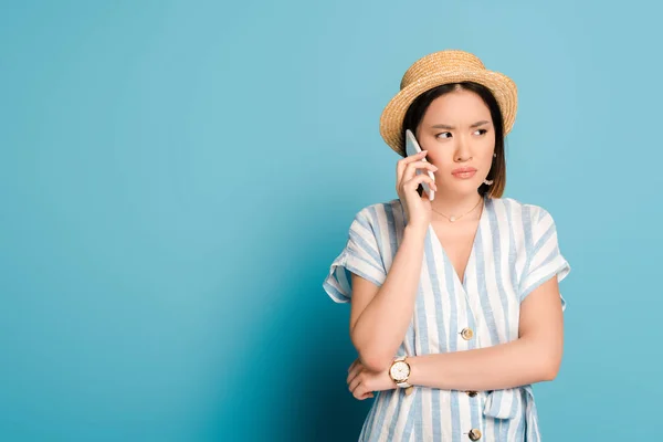 Triste morena asiática chica en rayas vestido y paja sombrero tomando en smartphone sobre fondo azul - foto de stock