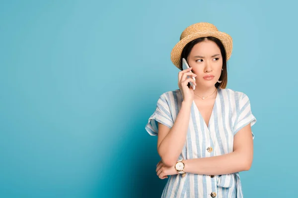 Triste morena asiática menina no vestido listrado e chapéu de palha tomando no smartphone no fundo azul — Fotografia de Stock