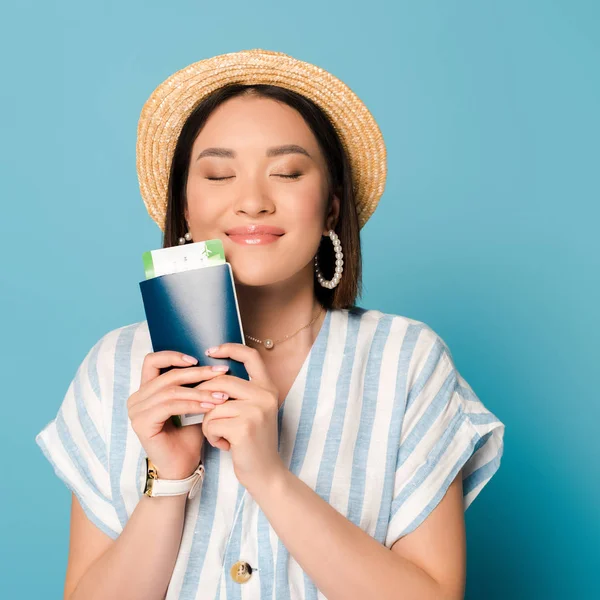 Erfreut brünette asiatische Mädchen in gestreiftem Kleid und Strohhut mit Pass mit Bordkarte auf blauem Hintergrund — Stockfoto