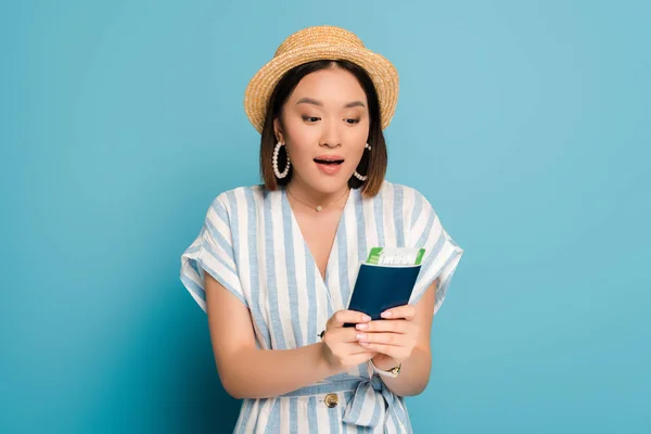 Excitada chica asiática morena en vestido a rayas y sombrero de paja con pasaporte con tarjeta de embarque sobre fondo azul - foto de stock