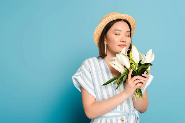 Sorridente ragazza asiatica bruna in abito a righe e cappello di paglia che tiene bouquet di gigli con gli occhi chiusi su sfondo blu — Foto stock
