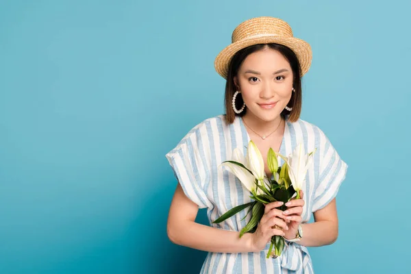 Sonriente morena asiática chica en vestido a rayas y sombrero de paja celebración ramo de lirios sobre fondo azul - foto de stock