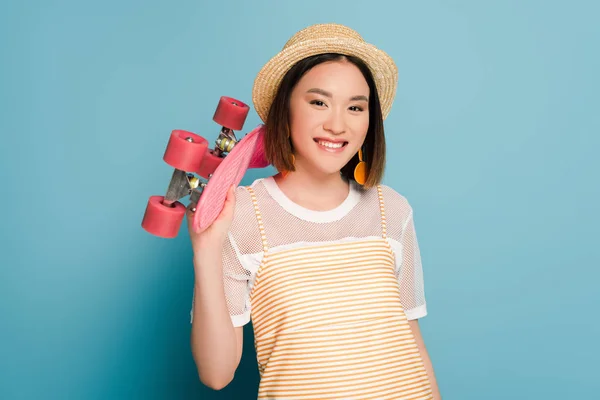 Sorridente ragazza asiatica in abito giallo a righe e cappello di paglia con penny board rosa su sfondo blu — Foto stock