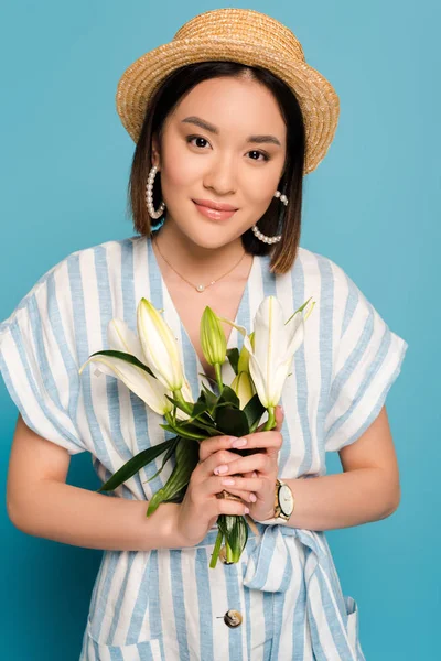 Morena chica asiática en vestido a rayas y sombrero de paja celebración ramo de lirios sobre fondo azul - foto de stock