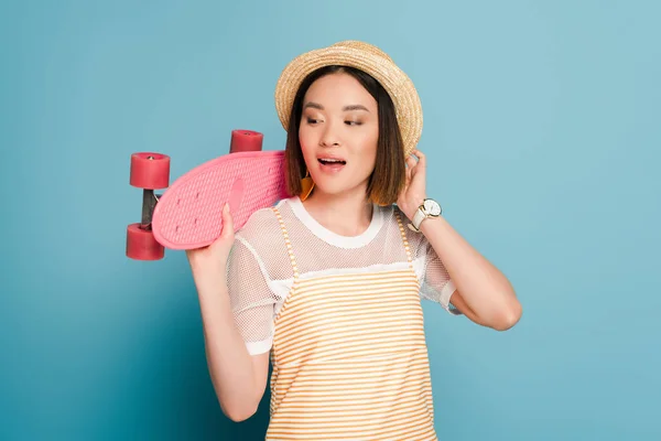 Asian girl in striped yellow dress and straw hat with pink penny board on blue background — Stock Photo