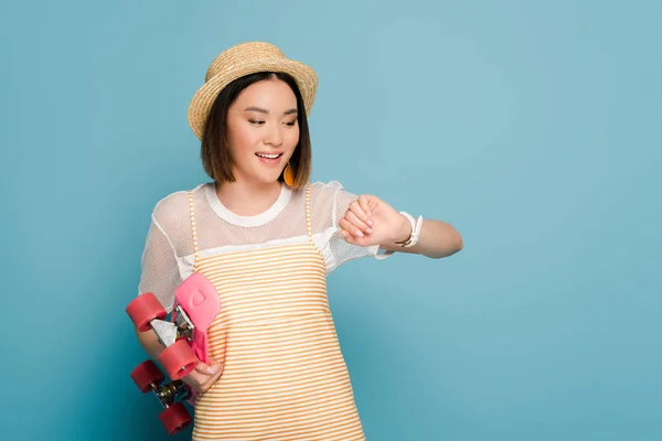 Smiling asian girl in striped yellow dress and straw hat with pink penny board looking at wristwatch on blue background — Stock Photo