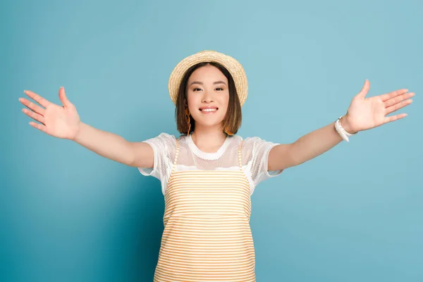 Sonriente chica asiática en rayas vestido amarillo y sombrero de paja con los brazos abiertos sobre fondo azul - foto de stock