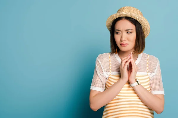 Skeptical asian girl in striped yellow dress and straw hat looking away on blue background — Stock Photo
