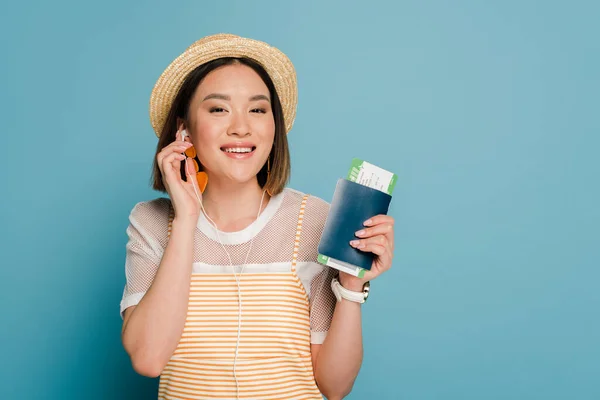 Sonriente asiático chica en rayas amarillo vestido y paja sombrero celebración pasaporte con tarjeta de embarque y hablando en teléfono inteligente sobre fondo azul - foto de stock