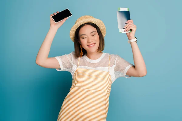 Sonriente asiático chica en rayas amarillo vestido y paja sombrero celebración pasaporte con tarjeta de embarque y teléfono inteligente sobre fondo azul - foto de stock