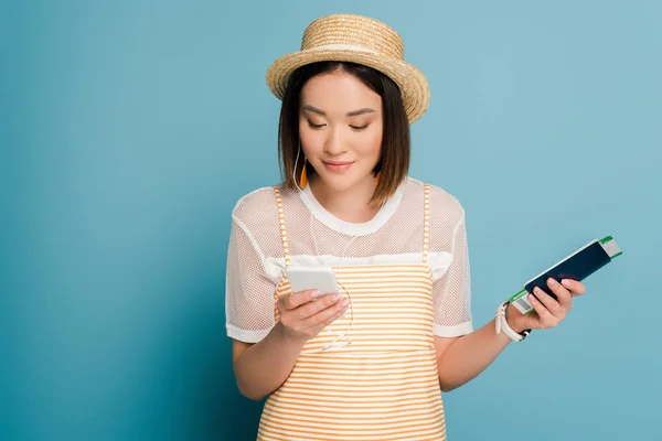 Smiling asian girl in striped yellow dress and straw hat holding passport with boarding pass and smartphone on blue background — Stock Photo