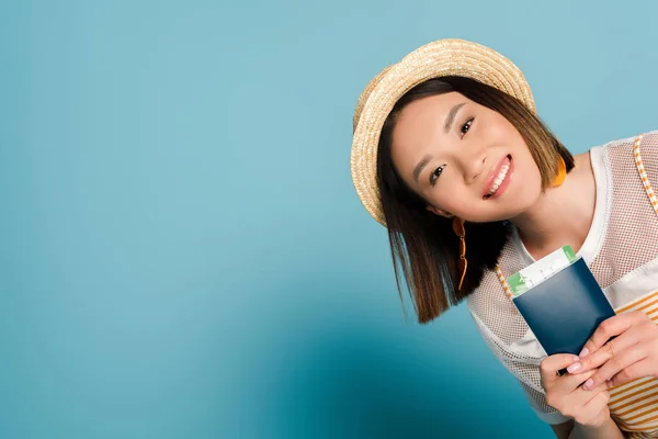 Sonriente asiático chica en rayas amarillo vestido y paja sombrero celebración pasaporte con tarjeta de embarque en azul fondo - foto de stock