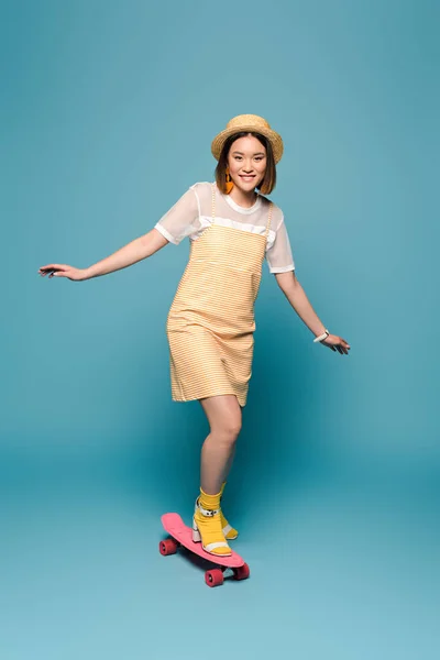Sonriente asiático chica en rayas amarillo vestido y paja sombrero cabalgando penny board en azul fondo - foto de stock