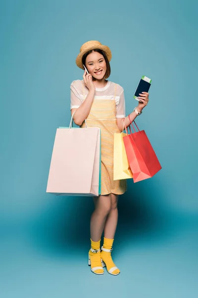 Sourire asiatique fille en robe jaune rayé et chapeau de paille avec des sacs à provisions et passeport parler sur smartphone sur fond bleu — Photo de stock