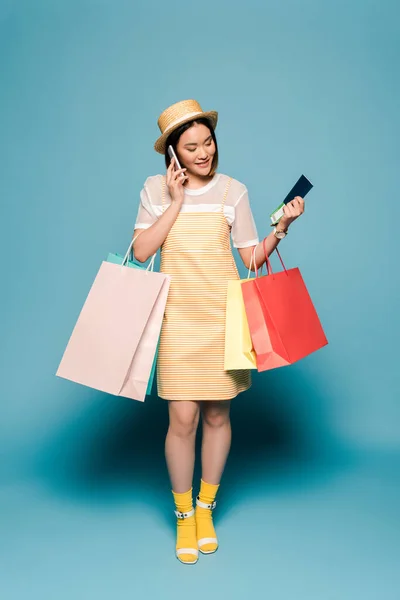 Lächelndes asiatisches Mädchen in gestreiftem gelben Kleid und Strohhut mit Einkaufstaschen und Reisepass im Gespräch auf Smartphone auf blauem Hintergrund — Stockfoto