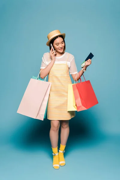 Sonriente chica asiática en rayas vestido amarillo y sombrero de paja con bolsas de compras y pasaporte hablando en el teléfono inteligente sobre fondo azul - foto de stock