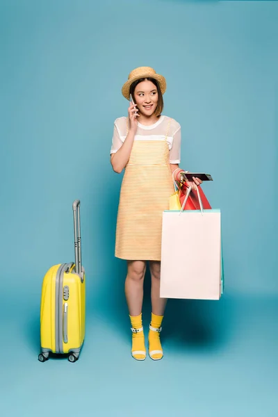Sorridente ragazza asiatica in abito giallo a righe e cappello di paglia con shopping bags, passaporto e valigia parlando su smartphone su sfondo blu — Foto stock