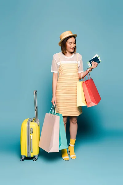 Sorridente ragazza asiatica in abito giallo a righe e cappello di paglia con borse della spesa, passaporto e valigia su sfondo blu — Foto stock