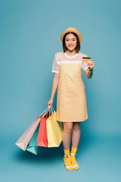 Sonriente chica asiática en rayas vestido amarillo y sombrero de paja con bolsas de compras y tarjeta de crédito sobre fondo azul - foto de stock