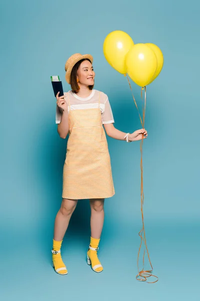 Sourire asiatique fille en robe rayée et chapeau de paille avec des ballons jaunes et passeport sur fond bleu — Photo de stock