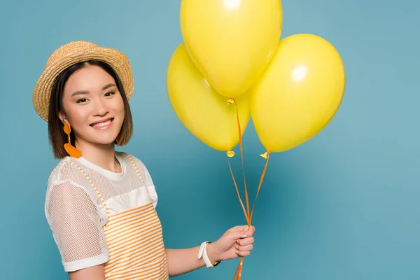 Feliz menina asiática em vestido listrado e chapéu de palha com balões amarelos no fundo azul — Fotografia de Stock