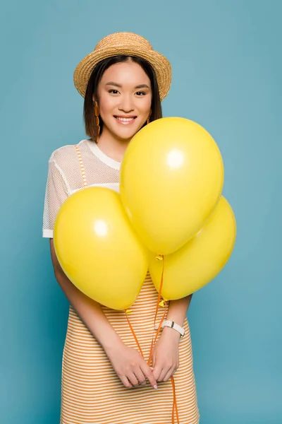 Feliz menina asiática em vestido listrado e chapéu de palha com balões amarelos no fundo azul — Fotografia de Stock