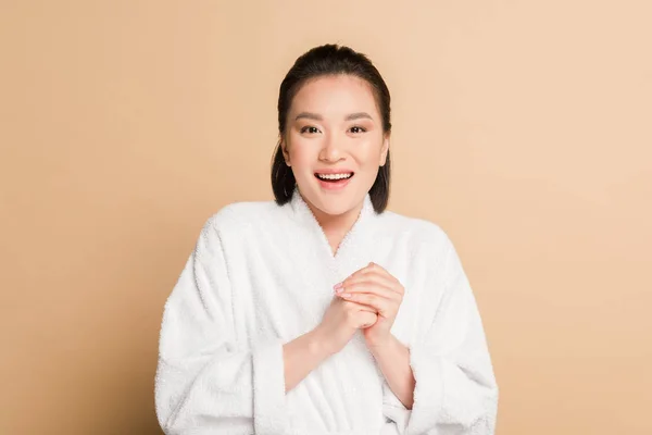 Happy beautiful asian woman in bathrobe touching face on beige background — Stock Photo