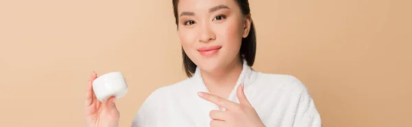 Sonriente hermosa mujer asiática en albornoz apuntando con el dedo a la crema cosmética aislado en beige, tiro panorámico - foto de stock