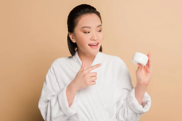 Sonriente hermosa mujer asiática en albornoz apuntando con el dedo a la crema cosmética sobre fondo beige - foto de stock
