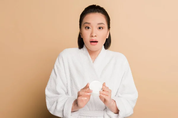 Shocked beautiful asian woman in bathrobe holding cosmetic cream on beige background — Stock Photo