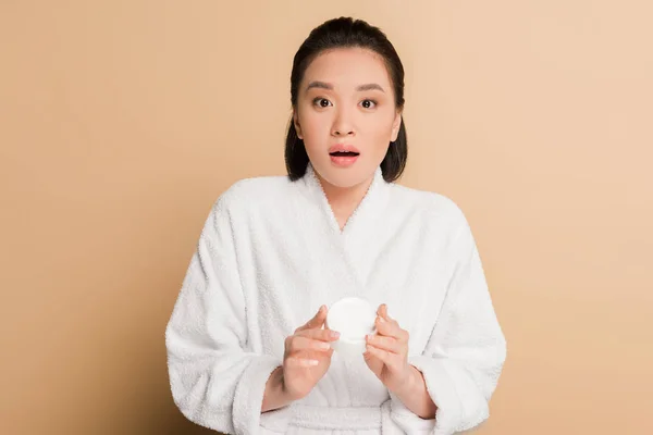 Schockiert schöne asiatische Frau im Bademantel hält kosmetische Creme auf beigem Hintergrund — Stockfoto