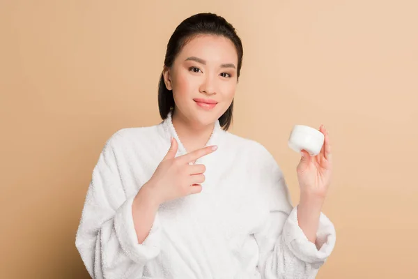 Sonriente hermosa mujer asiática en albornoz apuntando con el dedo a la crema cosmética sobre fondo beige - foto de stock