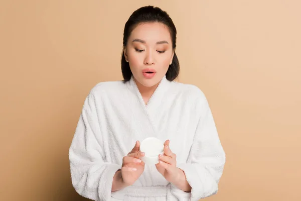 Shocked beautiful asian woman in bathrobe holding cosmetic cream on beige background — Stock Photo