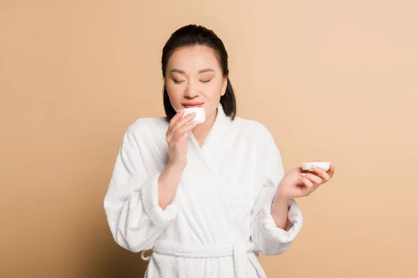 Smiling beautiful asian woman in bathrobe smelling cosmetic cream on beige background — Stock Photo