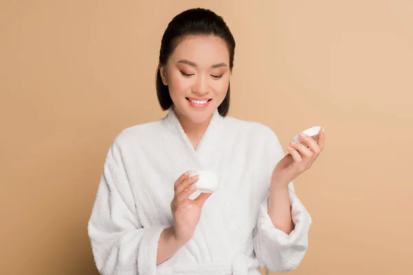 Smiling beautiful asian woman in bathrobe holding cosmetic cream on beige background — Stock Photo