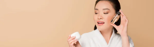 Smiling beautiful asian woman in bathrobe holding cosmetic cream and oil isolated on beige, panoramic shot — Stock Photo