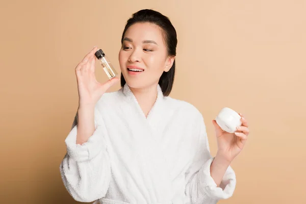 Smiling beautiful asian woman in bathrobe holding cosmetic cream and oil on beige background — Stock Photo